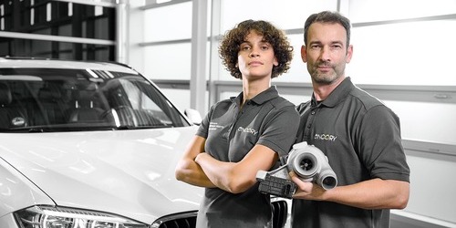 ZweiWerkstattmitarbeitendeinEncoryUniformstehenvoreinemweienBMWeinePersonhlteinwiederaufbereitetesErsatzteilindenHnden..jpg